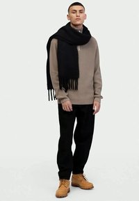 Young man wearing beige sweater, black scarf draped over shoulders, black pants, and tan boots, standing against a plain light background.