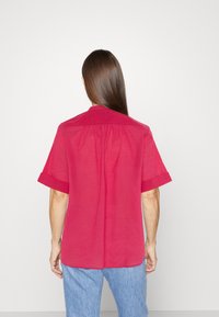Maglietta rosa brillante a maniche corte con colletto a punta, pieghe posteriori e dettagli delle maniche arrotolate, indossata con jeans blu chiaro.