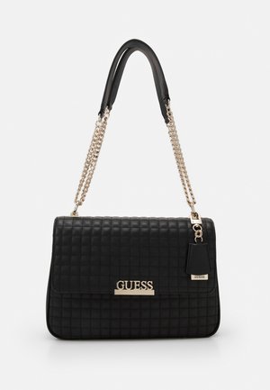 Czarna pikowana torebka na złotym łańcuszkowym pasku, z złotą blachą z logo "GUESS" oraz dopasowaną zawieszką, przedstawiona na jednolitym białym tle.