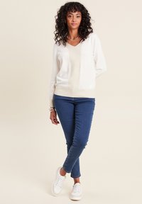 Maglione chiaro con scollo a V e pannello testurizzato a contrasto, abbinato a jeans blu slim-fit e scarpe da ginnastica bianche.