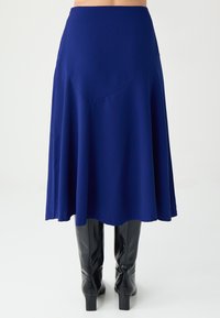 Jupe évasée taille haute bleu marine en tissu lisse, avec une silhouette fluide et des coutures angulaires discrètes. Portée avec des bottes noires montantes jusqu'au genou.