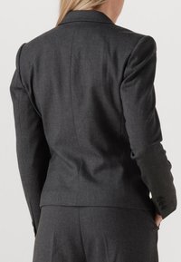 Kvinna i en skräddarsydd mörkgrå kavaj och matchande byxor, poserande med händerna i fickorna, bak- och sida synliga, mot en ljus, enfärgad bakgrund.