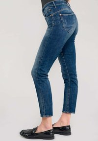 Jean en denim bleu skinny avec une taille mi-haute, des accents décolorés, des accessoires en cuivre et cinq poches ; associés à des mocassins noirs en vernis.
