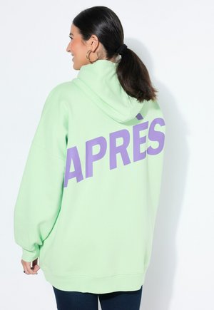 Kvinde med mørkt hår i hestehale bærer en lysegrøn hættetrøje med stor lilla tekst "APRÈS" på ryggen, kombineret med mørke bukser.