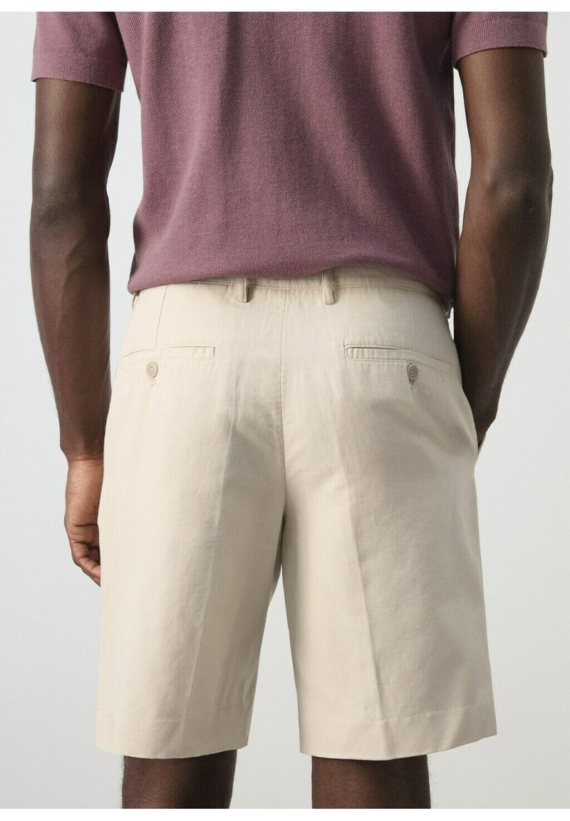 Homme portant un short beige mi-long avec des poches arrière à boutons et un pull à manches courtes mauve, vu de dos.