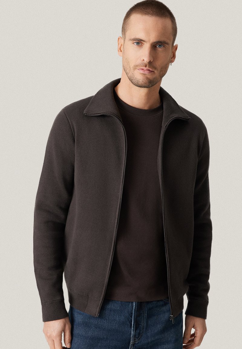 Mężczyzna z krótkimi włosami i brodą, ubrany w ciemnobrązowy sweter z zamkiem i dżinsy, stojący na tle jasnego, jednolitego tła.