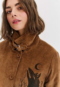 Brązowa kurtka z korduroy z wysokim kołnierzem, z grafiką wilka, guzikami i teksturowanym materiałem. Subtelny połysk dodaje głębi.