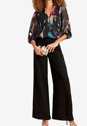 Kvinde iført en sort blomstret bluse, brede sorte bukser og holder en geometrisk metallisk clutch, med krøllet hår og sandaletter med remme.