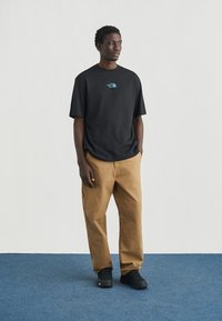 Μαύρο κοντομάνικο μπλουζάκι με μπλε λογότυπο, σετ με καφέ άνετο παντελόνι και μαύρα αθλητικά παπούτσια, στέκεται σε μπλε χαλί.