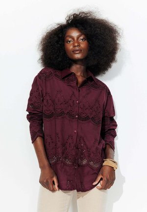 Chemise boutonnée bordeaux avec des détails brodés et des motifs découpés, à manches longues et à coupe décontractée. Le modèle porte des bracelets.