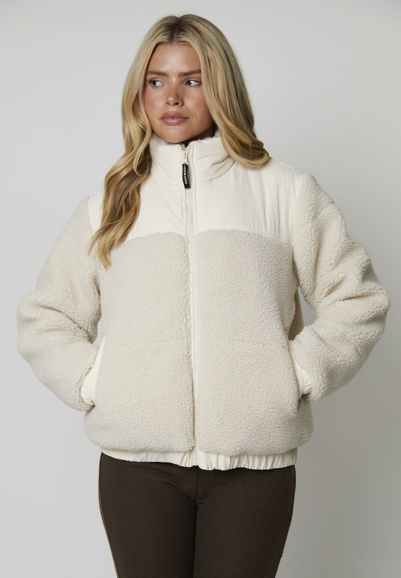 Mujer con cabello largo y rubio, vistiendo una chaqueta de forro polar sherpa color crema con bolsillos laterales y cuello con cremallera, mirando hacia su izquierda.