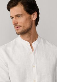 Hombre con cabello corto castaño y barba, vestido con una camisa blanca de lino con cuello mandarín, mirando hacia la izquierda sobre un fondo neutro.