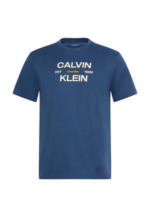 Blå t-skjorte med korte ermer med "CALVIN KLEIN" trykt i store beige bokstaver og "EST 1968" med en liten Calvin Klein-etikett i midten.