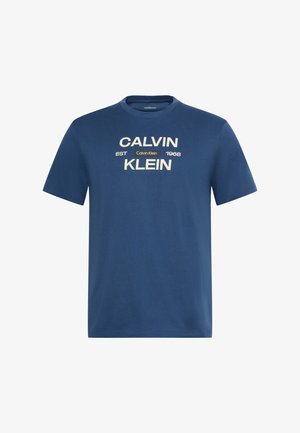 Μπλε κοντομάνικη μπλούζα με το «CALVIN KLEIN» τυπωμένο με μεγάλα μπεζ γράμματα και «EST 1968» με μια μικρή ετικέτα Calvin Klein στο κέντρο.