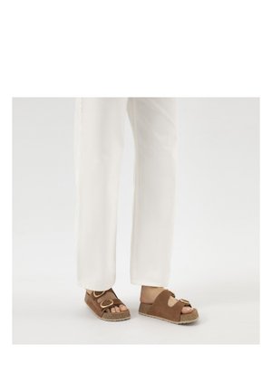 Personne portant un pantalon blanc et des sandales en liège marron à boucles dorées, debout devant un fond clair et uni.