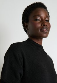 Pull en maille noire avec un col rond côtelé, un tissu texturé et une coupe décontractée. Cheveux courts et bouclés du modèle visibles.