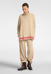 Maglione beige a collo alto lavorato a maglia con strisce rosse orizzontali all'orlo, abbinato a pantaloni beige con strisce rosse laterali e stivali alla caviglia marroni.