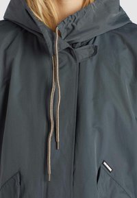 Wodoodporna ciemnoszara kurtka z kapturem ze sznurkiem, beżowymi akcentami w postaci sznurków oraz boczną kieszenią. Ma małą metkę z logo na dolnym brzegu.