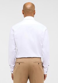 Weißes Langarmhemd mit klassischem Kragen, kombiniert mit beigen Hosen, die zwei Gesäßtaschen und Knopfakzente haben.