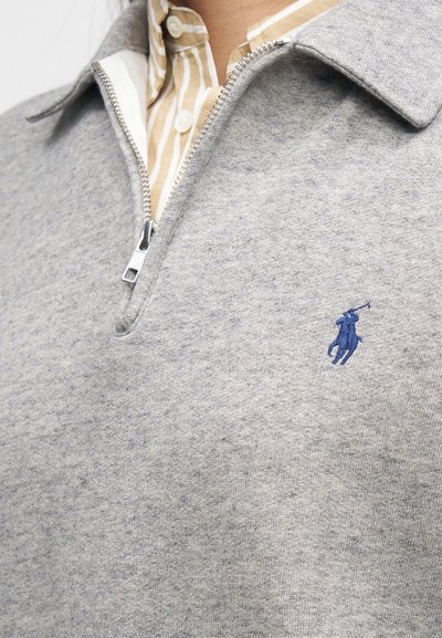 Siv pulover z zadrgo iz mehke tkanine, ki ima ovratnik in majhen moder vezen logo na prsih. Pod njim je vidna črtasta majica.