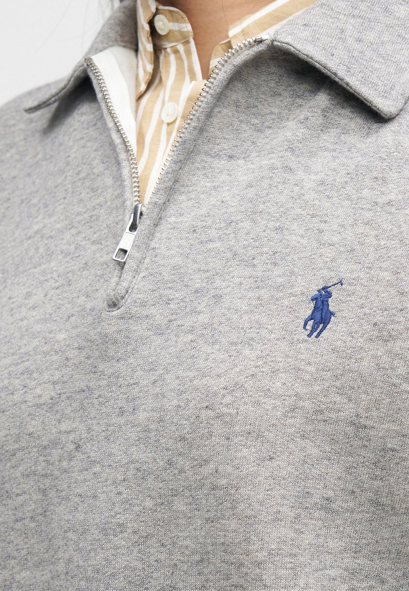 Pull gris à fermeture éclair en tissu doux, avec un col et un petit logo bleu brodé sur la poitrine. En dessous, une chemise rayée est visible.