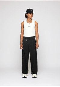 Camiseta branca sem mangas, calças largas pretas com passantes para cinto, detalhes em metal prateado e ténis creme. Boné preto completa o visual.