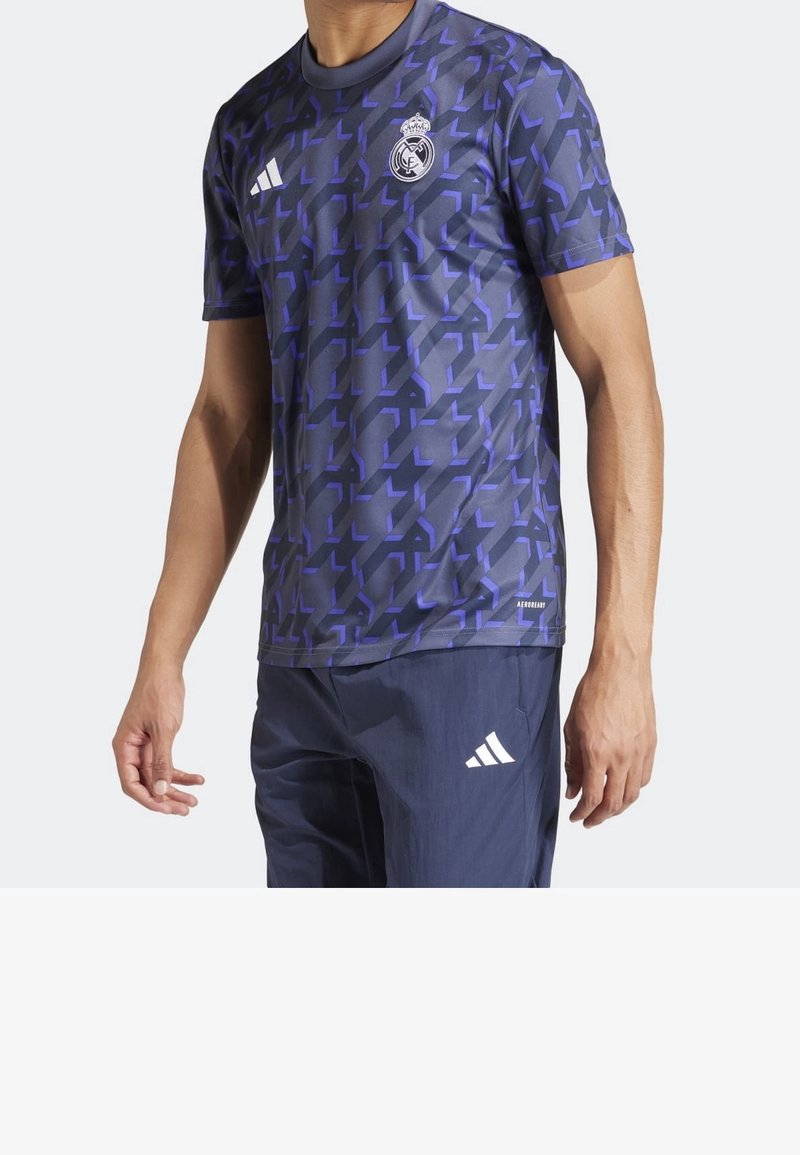 Muž v modro-černém geometrickém vzoru dresu Real Madrid s logem Adidas a tmavě modrých kalhotách Adidas.