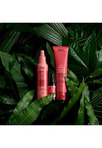 Trois produits capillaires Aveda Nutri Plenish dans des nuances de rose : un spray, une bouteille avec pompe et un tube, posés sur des feuilles vertes texturées.