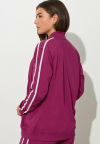 Joven mujer con una chaqueta de chándal magenta con franjas blancas en las mangas, de perfil a la izquierda, con las manos en los bolsillos de la chaqueta, frente a un fondo liso.