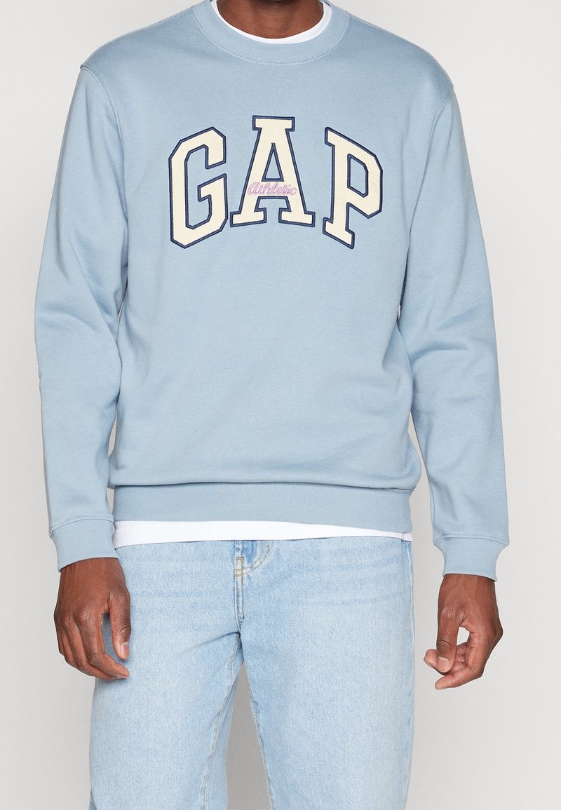 Homme portant un sweat-shirt bleu clair avec de grandes lettres GAP et le mot Athletic en plus petit, associé à un jean bleu clair.