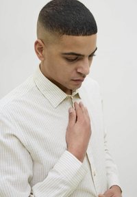Camisa de manga larga en color crema con rayas amarillas claras, cuello abotonado y una tela texturizada. El modelo toca la camisa con una mano.