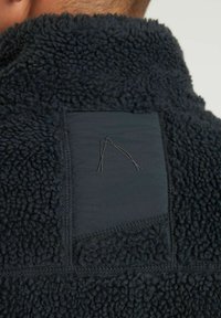 Giacca in pile nera con superficie morbida e testurizzata e un pannello in tessuto liscio a contrasto, dotata di un design ricamato sul retro.
