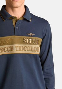 Polo a maniche lunghe blu navy con striscia orizzontale marrone chiaro, testo ricamato, dettagli dorati e un logo con ali sul petto.