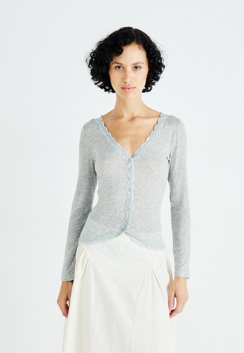Lichtgrijze langwerpige cardigan met geschulpte kantafwerking en knoopsluiting. Zacht stof met textuur ribbeling, gecombineerd met een witte rok.