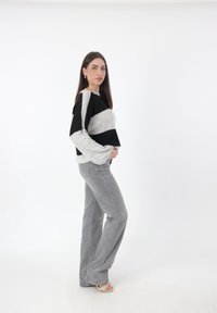 Schwarzer und grauer gestreifter Pullover mit weiten Ärmeln, kombiniert mit hellgrauen, ausladenden Jeans. Der Pullover hat ein strukturiertes Strickmuster; das Modell steht vor einem weißen Hintergrund.