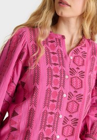 Blusa rosa con bottoni, caratterizzata da intricati motivi scuri, in un tessuto testurizzato e con un collo rotondo. Dettagli notevoli includono cuciture decorative.