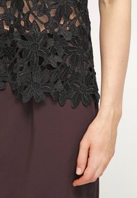 Haut en dentelle florale noire avec des pétales transparents porté sur une jupe violet foncé, montrant un gros plan d'une main détendue le long du corps.