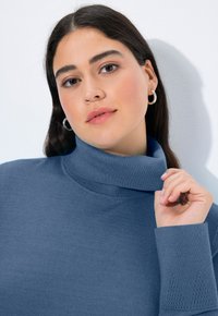 Vaaleansininen pooloneule, jonka pehmeä neulos sisältää ribbauskaitaleet hihoissa ja kauluksessa, sekä hienovaraisen teksturoidun yksityiskohdan hihassa.