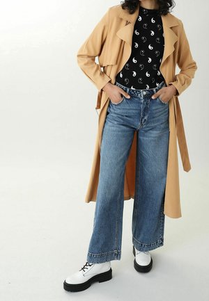 Femme portant un trench-coat beige, une chemise noire avec des motifs yin-yang blancs, un jean large bleu et des chaussures blanches à lacets, debout les mains dans les poches.