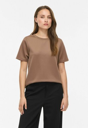 Jeune femme aux longs cheveux bruns portant un t-shirt marron uni à manches courtes et un pantalon noir sur fond blanc.