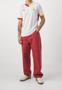 Vit v-ringad tröja med röda och gula detaljer, med en logotyp och tre stjärnor. Kombinerad med röda jeans och vita sneakers.