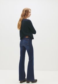 Hosszú világosbarna hajú nő sötét pulóverben, sötétkék harangnadrágban és fekete magassarkú cipőben áll, egy sima fehér háttér előtt.