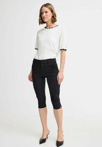 Weißes Kurzarmoberteil mit einem schwarzen Wellenhals, kombiniert mit enganliegenden schwarzen Caprihosen. Das Model trägt spitze schwarze High Heels und zeigt klare Linien.