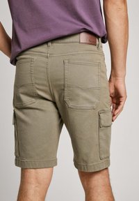 Khaki cargo šortky s prehnutými manžetami, dvoma zadnými vreckami a jedným bočným vreckom, vyrobené z odolnej bavlnenej tkaniny, s hnedou koženou etiketou.