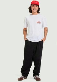 Camiseta branca de manga curta com um logotipo vermelho, combinada com calças largas pretas. Um boné vermelho e sapatilhas castanhas completam o look.