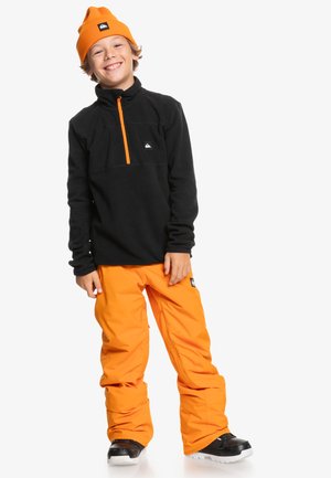 Pull-over en polaire noir avec fermeture éclair et accents orange, associé à un pantalon de neige orange ample. Des bottes noires complètent la tenue.
