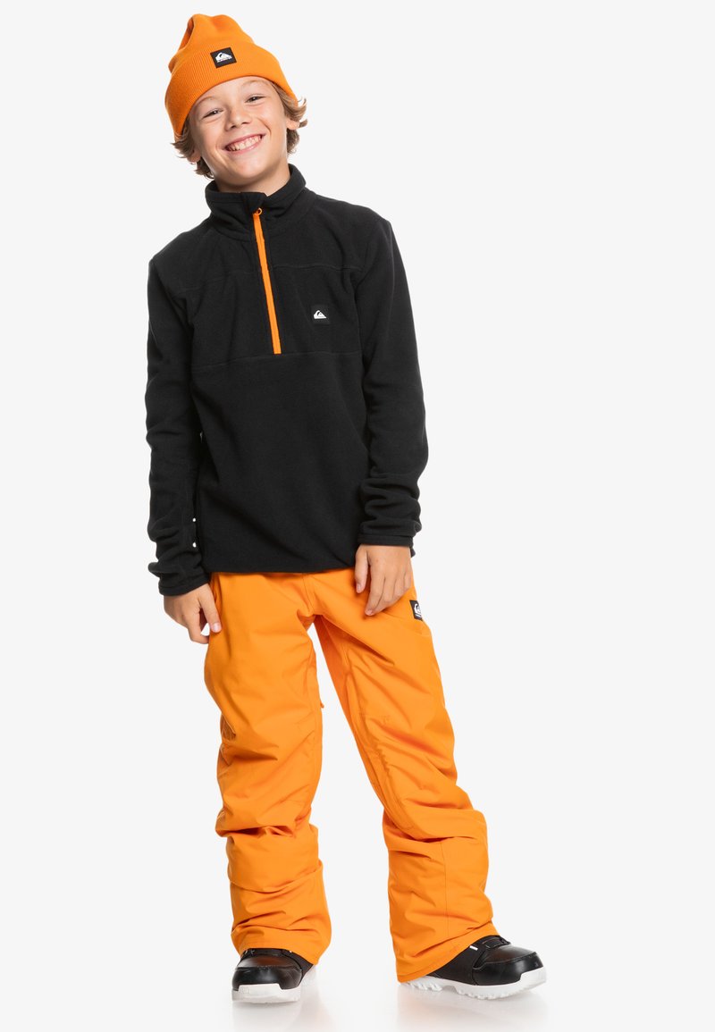 Schwarzer Fleece-Pullover mit orangefarbendem Reißverschluss und Akzenten, kombiniert mit lockeren orangen Schneehosen. Schwarze Stiefel runden das Outfit ab.