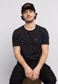 Zwarte T-shirt met een geometrisch patroon in grijs, met een klein oranje berenlogo op de borst. Model draagt een zwarte pet met een berenontwerp.