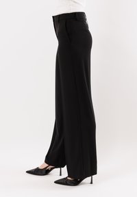 Pantaloni neri a gamba larga con una texture liscia, caratterizzati da un taglio sartoriale e dettagli plissettati. Indossati con tacchi neri a punta.