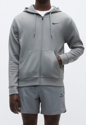 Mann trägt hellgrauen Nike-Zip-Hoodie und passende Shorts, eine Hand in der Kapuzentasche, steht vor einfachem Hintergrund.